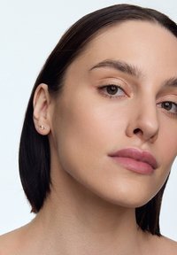 Złote kolczyki w kształcie małych kwiatów z delikatnymi detalami. Modelka ma ciemne włosy i naturalny makijaż, prezentując kolczyki.