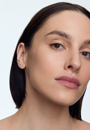 Złote kolczyki w kształcie małych kwiatów z delikatnymi detalami. Modelka ma ciemne włosy i naturalny makijaż, prezentując kolczyki.