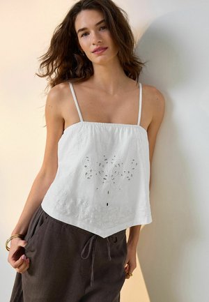 Femme aux cheveux bruns lâchés, portant un top blanc brodé sans manches et un pantalon sombre à cordon, debout contre un mur clair.