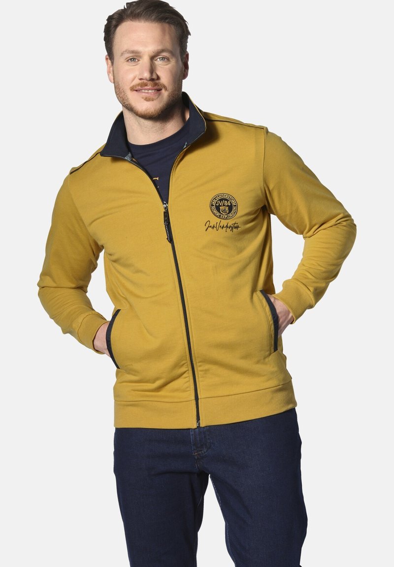 Gelbes Zip-Hoodie mit schwarzen Akzenten, gerippten Bündchen und einem runden Logo auf der Brust. Verfügt über Seitentaschen und einen hohen Kragen.