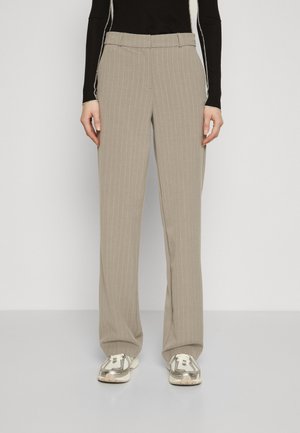 Persona vistiendo pantalones de pierna ancha con rayas beige y zapatillas blancas, de pie contra un fondo liso.