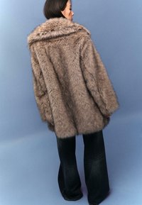 Cappotto in finta pelliccia marrone con ampio colletto e vestibilità morbida, caratterizzato da maniche lunghe e una superficie morbida e texturizzata. Abbinato a jeans svasati scuri.