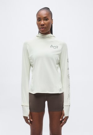 Γυναίκα που στέκεται στραμμένη προς τα εμπρός, φοράει ένα ανοιχτόχρωμο μακρυμάνικο με κουκούλα με το λογότυπο ACG και σκούρα στενά σορτς.