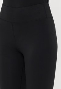 Juodos aukšto liemens priglundančios legging kelnės, matomos nuo liemens iki vidurinės šlaunies, su matomais siūlėmis ir lygiu audinio tekstūra.