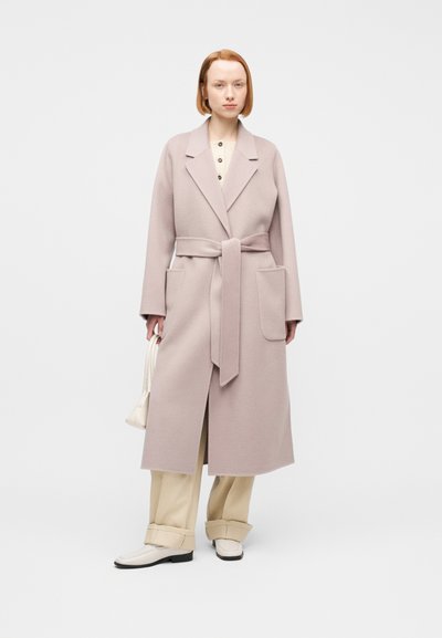 Γυναίκα με μακρύ μοβ παλτό με ζωνάκι, μπεζ πουκάμισο με κουμπιά και φαρδιά μπεζ παντελόνια, κρατώντας μικρή λευκή τσάντα, στέκεται σε λευκό φόντο.