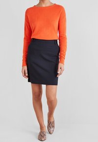 Pull orange vif à manches longues, jupe crayon bleu marine et mules beiges à imprimé serpent. Tissus lisses, coupe ajustée et silhouettes simples.