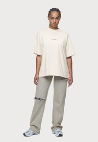 Beige suur suurusega t-särk punase "PEGADOR" tekstiga, koos heledate hallide teksadega, millel on kulunud detailid, ja sportlike hallide tossudega.