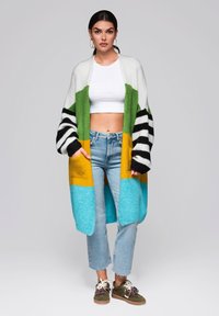 Yksinkertainen ylikokoinen neuletakki, jossa on vihreitä, keltaisia, sinisiä ja mustia raitoja; neulottu kangas; taskut; päällä valkoinen crop-top ja farkut.