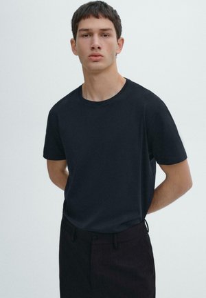 Young man standing with hands behind back wearing a plain black t-shirt and dark pants against a plain light background.