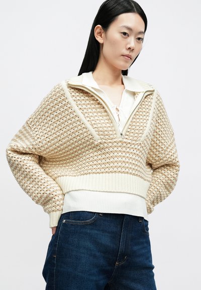 Béžový pletený pulóver s textúrovaným vzorom, s polopriehradkovým golierom a rebrovanými manžetami, kombinovaný s tmavomodrými džínsami.