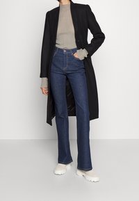Schwarzer Mantel mit strukturiertem Design, graues, geripptes Langarm-Oberteil, hochgeschnittene dunkelblaue Jeans und weiße Schuhe mit dickem Sohlen.