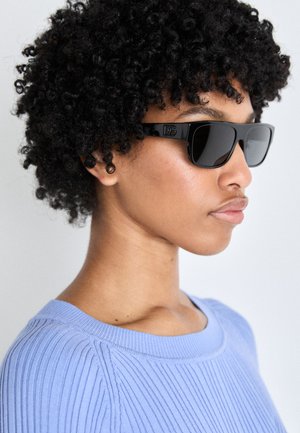 Occhiali da sole neri con lenti grandi e rettangolari e una montatura lucida, dotati di un logo discreto sulle aste. Indossati con un maglione a coste azzurro chiaro.