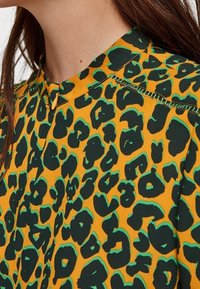 Chemise à imprimé léopard avec un fond jaune vif et des taches noires et vertes. Le col est structuré avec une patte de boutonnage minimaliste.