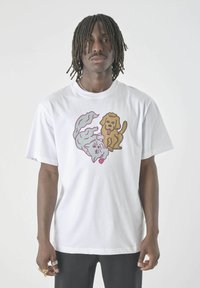 Mann trägt ein weißes T-Shirt mit einem grafischen Motiv von einer cartoonhaften Katze und einem Hund und steht vor einem schlichten hellen Hintergrund.