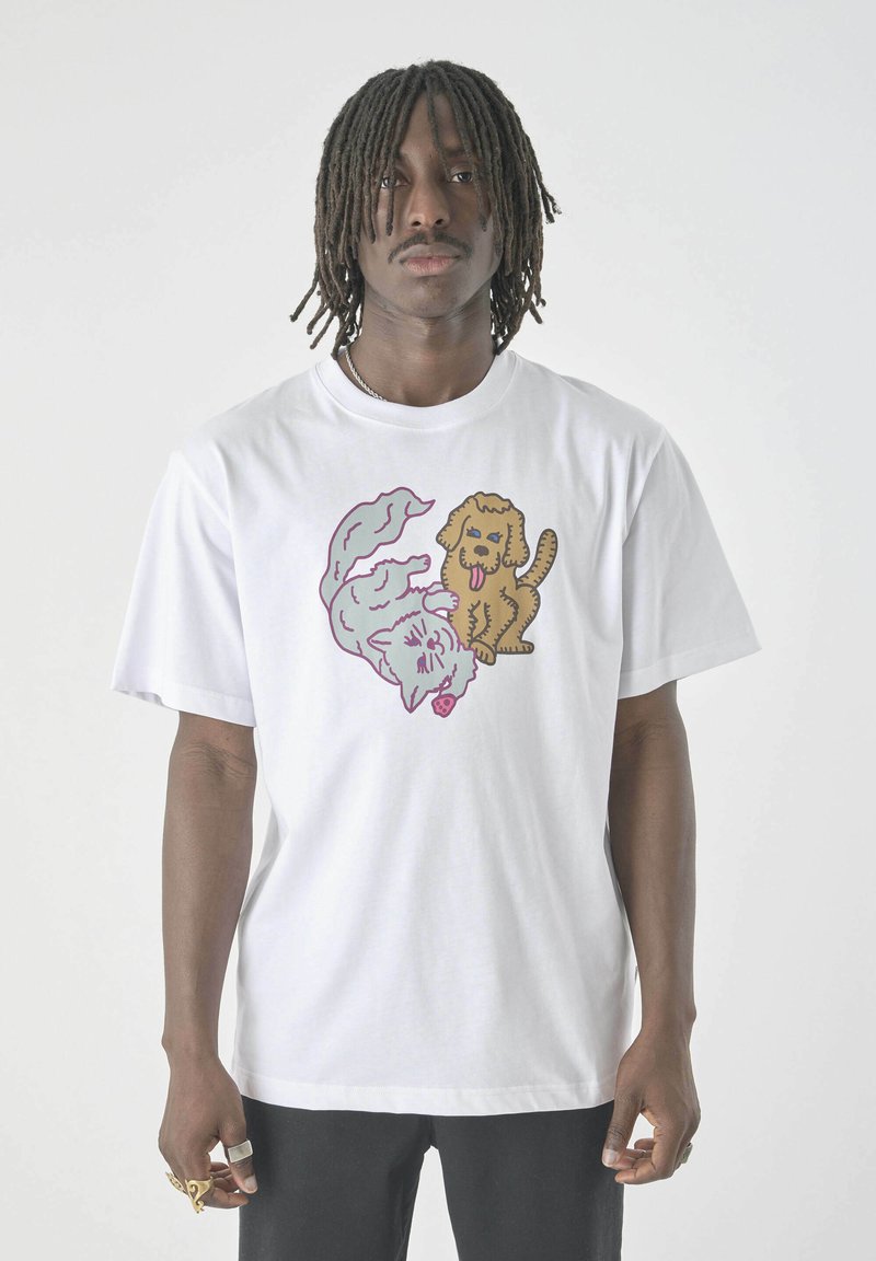 Mann trägt ein weißes T-Shirt mit einem grafischen Motiv von einer cartoonhaften Katze und einem Hund und steht vor einem schlichten hellen Hintergrund.