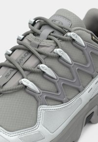 Γκρι αθλητικό παπούτσι με επάνω μέρος από δίχτυ, υφή με ανάγλυφα σχέδια, ασημένιες λεπτομέρειες και ανακλαστικά στοιχεία. Διαθέτει ασφαλές σύστημα κορδονιών και μαλακή σόλα.