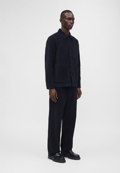 Homme debout portant une veste noire à boutons avec trois poches avant, un pantalon noir assorti et des chaussures noires sur un fond clair uni.