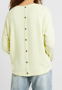 Jasnożółty dzianinowy sweter z okrągłym dekoltem, opuszczanymi ramionami i rzędem brązowych guzików na plecach. Widoczna miękka tekstura.