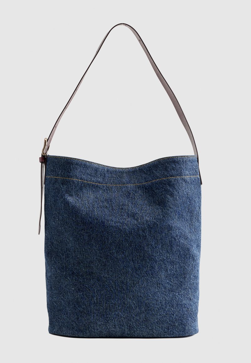 Blaue Jeans-Stofftasche mit einem verstellbaren braunen Schulterriemen und sichtbaren gelben Nähten vor einfarbigem Hintergrund.