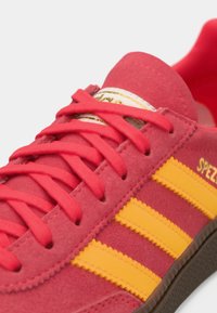 Κόκκινο σουέτ αθλητικό παπούτσι με τρεις κίτρινες ρίγες και κορδόνια. Διαθέτει υφασμένη λαστιχένια σόλα και ετικέτα γλώσσας με χρυσές λεπτομέρειες.