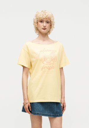 Ung person med krøllet blondt hår iført en gul T-shirt med teksten "always fresh" og et tekrus-grafik, kombineret med et blåt denimskørt.