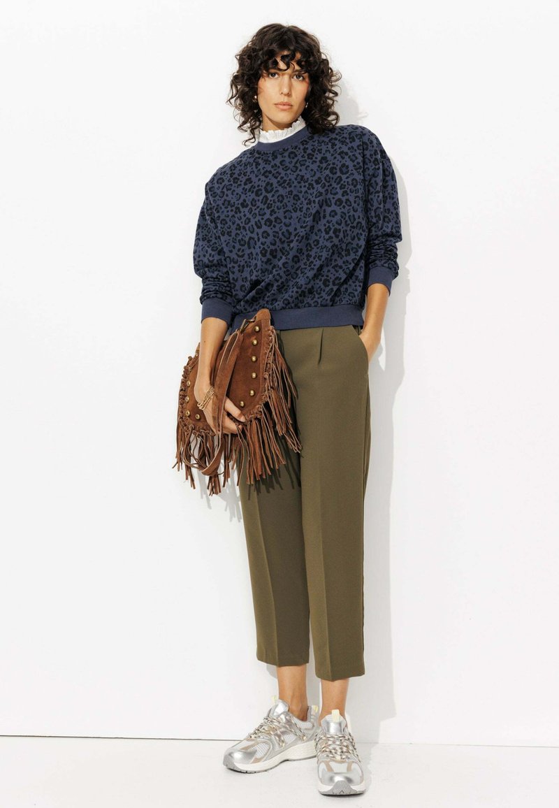 Sweatshirt en léopard navy, pantalons olive coupés, sac en franges marron avec clous, et baskets argentées avec accents jaunes.