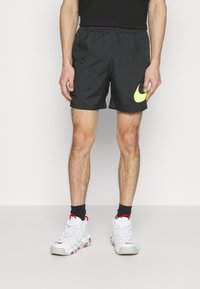 Man som bär svarta träningsshorts med neonröd Nike-logotyp, svarta strumpor och vita Nike-sneakers med röda detaljer, som står på en grå golv.
