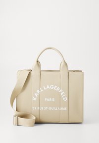 Sac fourre-tout texturé beige avec de courtes poignées et une bandoulière amovible. Présente un texte imprimé en blanc : "KARL LAGERFELD PARIS 21 RUE ST-GUILLAUME."
