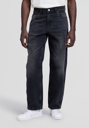 Person, die lockere schwarze Jeans mit einem verblassten Effekt und weißen Sneakers trägt, steht vor einem hellgrauen Hintergrund.