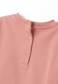 Top de algodón rosa claro con cuello redondo, collar acanalado y cierres a presión en la parte delantera para facilitar su uso. Textura suave.