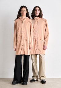 Peach-colored raincoats with hoods, featuring snap buttons, front pockets, and a relaxed fit, worn over dark and light trousers with chunky shoes.