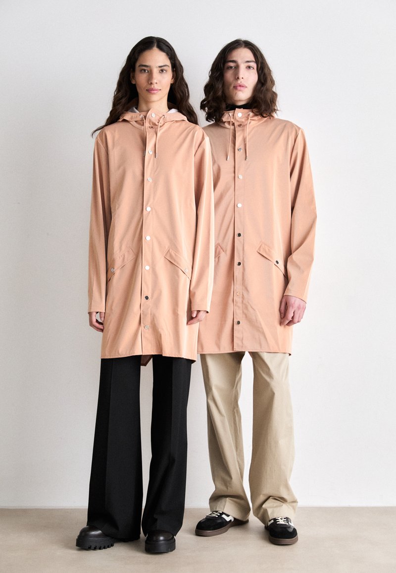 Peach-colored raincoats with hoods, featuring snap buttons, front pockets, and a relaxed fit, worn over dark and light trousers with chunky shoes.