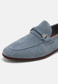 Mocassin en daim bleu présentant une forme plate, une bande accentuée tressée et un détail boucle en ton argenté. Semelle extérieure marron contrastant avec la tige.
