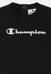 Svart bomulls t-skjorte med rund hals, med en hvit "Champion"-logo på brystet og med trykknappåpning bak. Glatt tekstur.