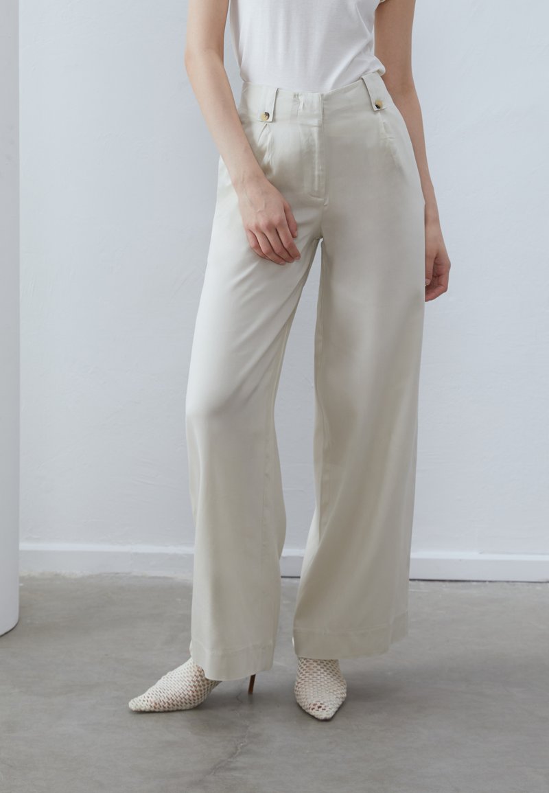 Crème-kleurige wijde broek van lichtgewicht materiaal, met voorzakken en goudkleurige knopen, gecombineerd met witte hakken met textuur.