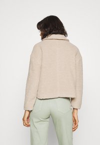 Beżowa krótka kurtka z miękkiego, teksturowanego materiału z kołnierzem, noszona na jasnoszare spodnie. Posiada długie rękawy i luźny krój.