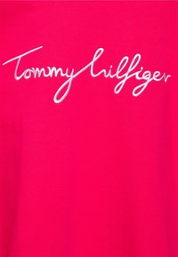 T-shirt di cotone rosa shocking con logo "Tommy Hilfiger" ricamato in bianco in font corsivo, posizionato su uno sfondo liscio e texturizzato.