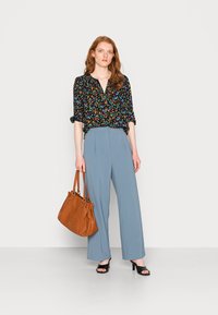 Blusa preta com flores e mangas dobradas, calças largas de tom azul claro, sapatos de salto abertos pretos e uma mala de ombro castanha.