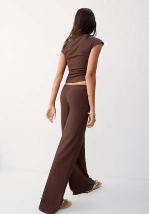 Femme marchant de dos portant un haut marron sans manches et un pantalon à jambes larges avec des sandales, sur un fond blanc uni.