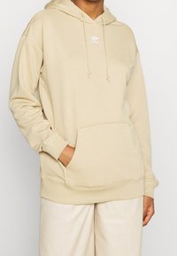 Sudadera con capucha beige hecha de tela suave, con bolsillo frontal, puños acanalados y un logo bordado en el pecho.