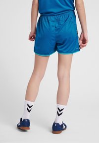 Osoba v modrých sportovních šortkách, bílých ponožkách s černými chevrony a modrých vnitřních sportovních botách, stojící na hladké světle šedé podlaze.