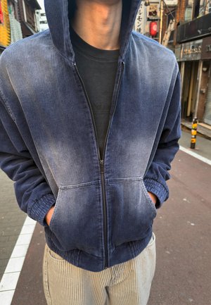 Egy személy kék kordbársony cipzáras kapucnis pulóvert visel, kezét a zsebében tartja, egy utcán áll, háttérben épületekkel és táblákkal.