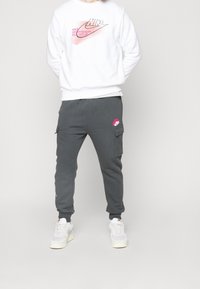 Vit sweatshirt med rosa Nike-logotyp och bokstäver, tillsammans med mörkgrå cargobyxor med fickor och en rosa accentlogotyp. Vita sneakers.