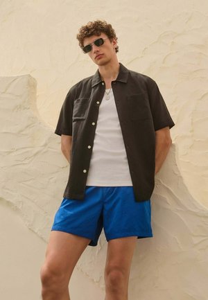 Junger Mann trägt schwarzes, kurzärmliges Hemd mit Knöpfen, weißes Tanktop, blaue Shorts und Sonnenbrille, steht vor einer strukturierten, beigen Wand.