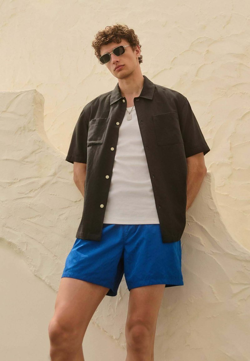 Jeune homme portant une chemise noire à manches courtes boutonnée, un débardeur blanc, un short bleu et des lunettes de soleil, debout devant un mur beige texturé.