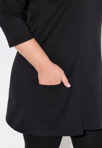 Tunika nera con maniche a tre quarti, dotata di una tasca laterale e di una texture liscia. L'orlo si trova sopra i leggings. Il tessuto appare leggero.