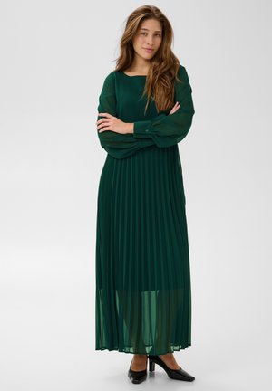 Une femme pose les bras croisés, vêtue d’une longue robe plissée vert émeraude et de talons noirs, sur un fond de studio gris clair.