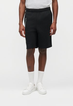 Férfi fekete térdig érő rövidnadrágban, fehér bokazokniban, fehér tornacipőben és fehér pólóban, egyszínű háttér előtt állva.