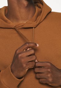 Felpa in cotone marrone con cappuccio e lacci. Vista ravvicinata di mani che regolano il colletto. Texture morbida e polsini a coste sono visibili.