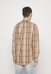 Camicia a quadri marrone con maniche lunghe, polsini con bottoni e un dettaglio sul dorso. Il tessuto presenta un motivo a scacchi in diverse tonalità di marrone e crema.